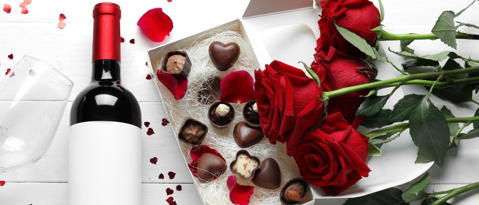 A bottle of red wine lying next to an open box of chocolate and a bouquet of roses